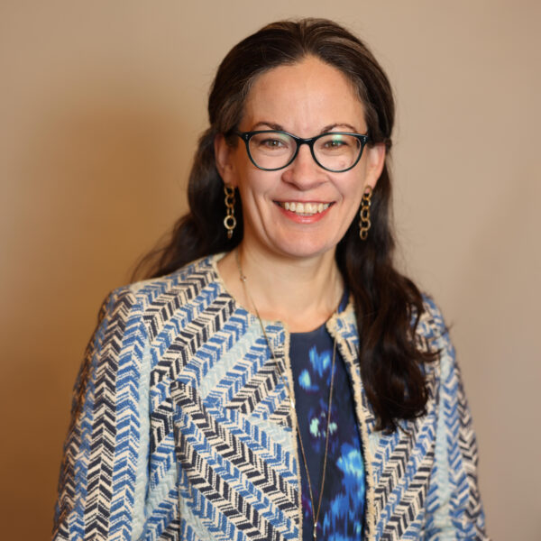 A woman with long dark hair and glasses smiles at the camera. She wears a blue patterned jacket over a dark blue top and gold earrings, standing with her hands resting on a chair against a neutral background.