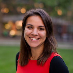A young woman with straight brown hair smiles at the camera. She is wearing a bright red top and standing outdoors, with a blurred green and brown background suggesting a park or campus setting.