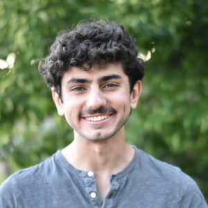 A young man with curly dark hair, wearing a gray buttoned shirt, smiles at the camera. Lush green trees form a blurred background, creating a natural outdoor setting.