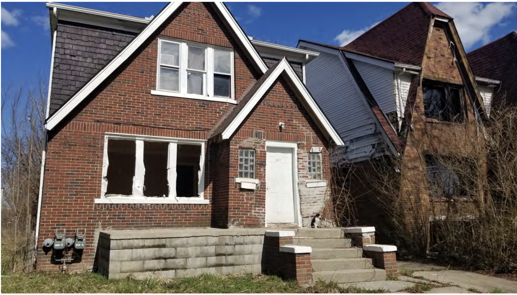 A vacant, red-brick house with broken windows and a damaged door sits on a concrete stoop, surrounded by overgrown weeds and next to another deteriorating home under a blue sky.