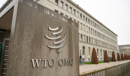 The exterior of the World Trade Organization (WTO) building in Geneva, Switzerland, featuring the WTO/OMC logo on a large stone sign in the foreground and the building with arched windows in the background.