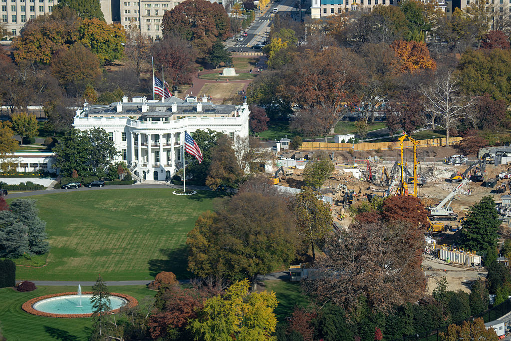 Aerial view of the White House surrounded by trees with autumn foliage. Construction work with heavy equipment is visible on the right side of the lawn. The American flag flies above the White House.