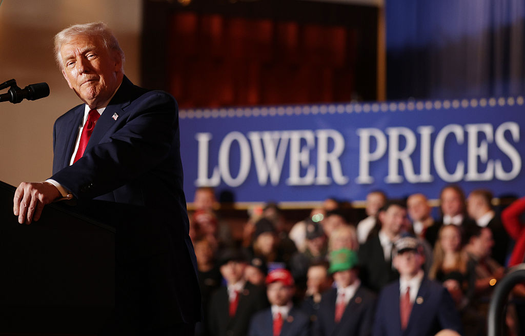 President Trump speaks at a podium with a crowd behind him and a large LOWER PRICES sign in the background.