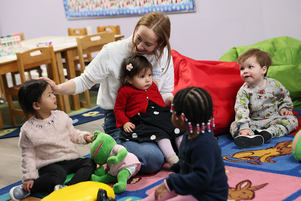 A woman sits on the floor in a playroom with four young children, engaging with them. The children are sitting on a colorful rug, surrounded by toys. Everyone appears happy and engaged in a playful activity.