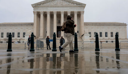 A person walks past the U.S. Supreme Court building on a rainy day, with their reflection visible in a wet pavement. Several other people stand near the entrance in the background.