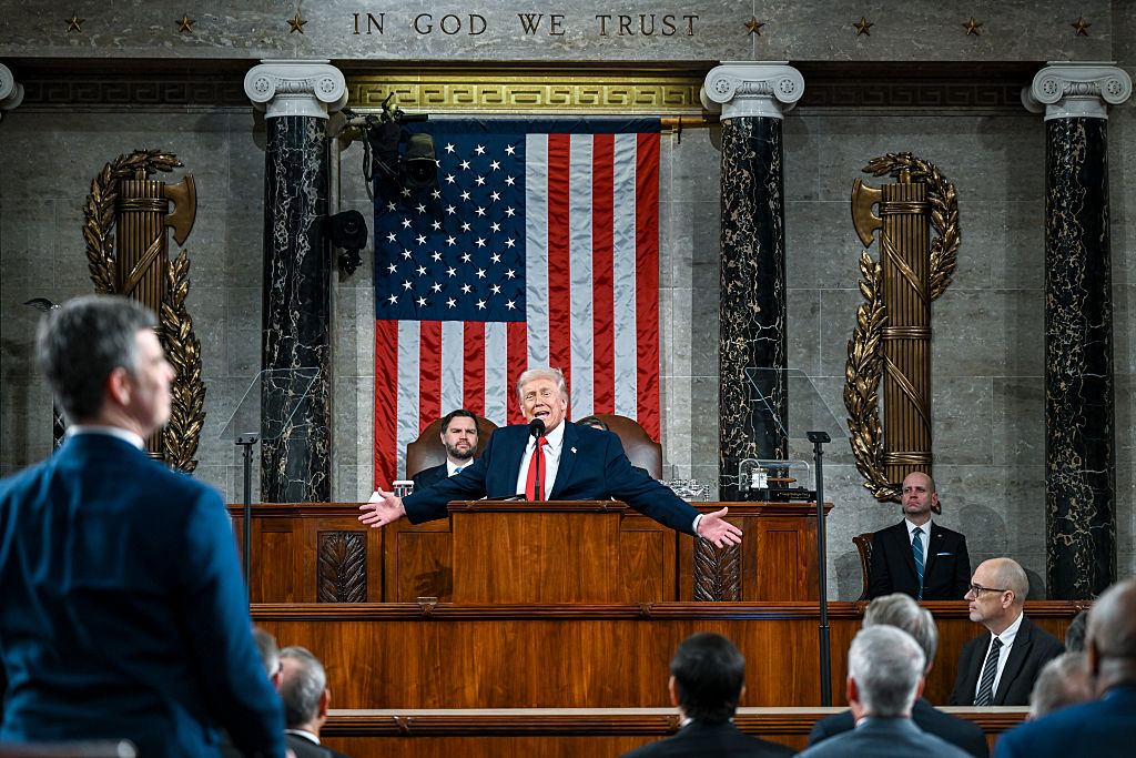 A man in a suit speaks passionately at a podium in a grand hall with an American flag behind him, surrounded by other officials and audience members. The words IN GOD WE TRUST are displayed above.