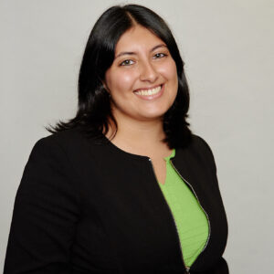 A woman with straight dark hair smiles at the camera. She is wearing a black blazer over a bright green top and stands against a plain light background.