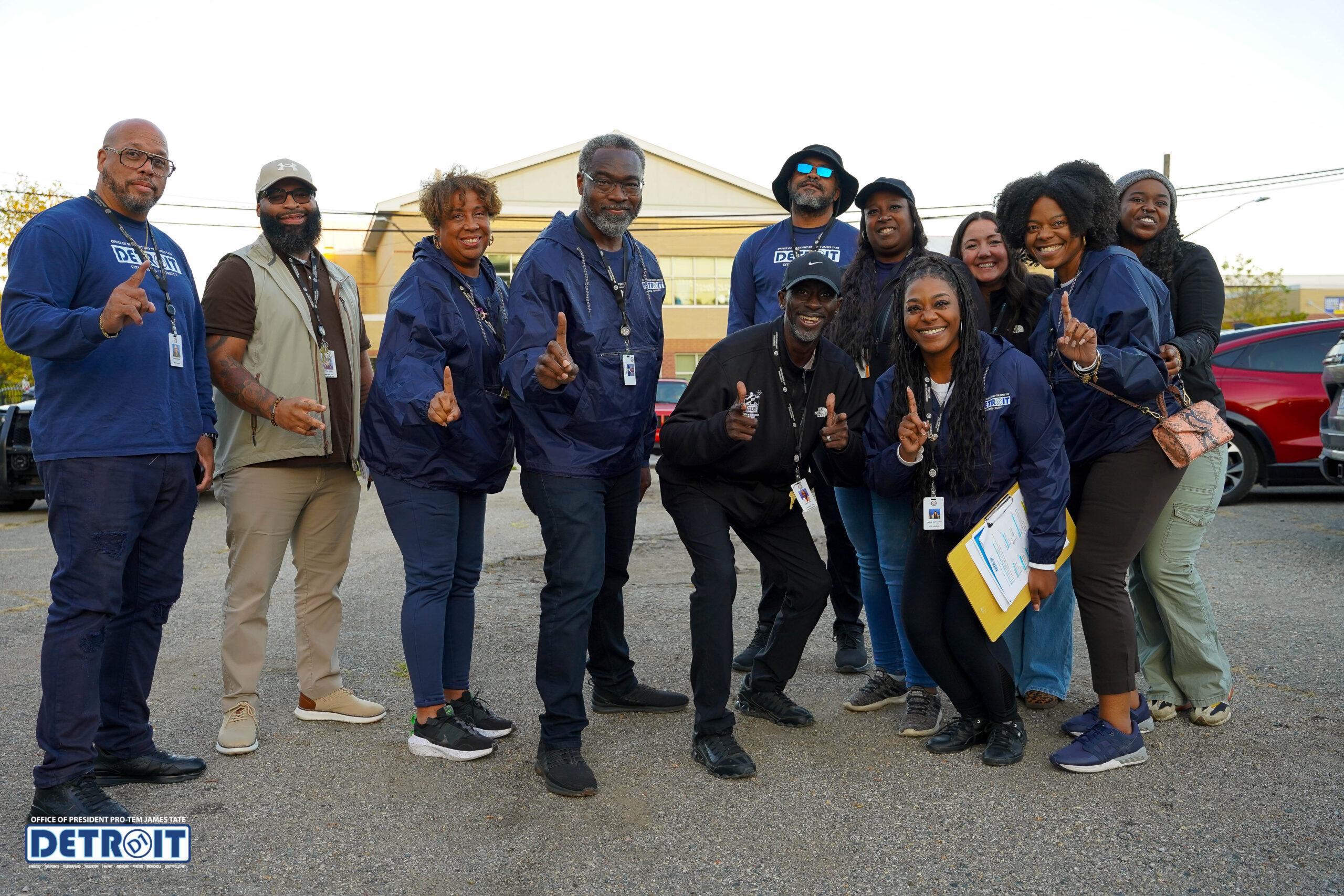 A group of twelve smiling adults pose together outdoors, many wearing navy blue jackets and badges. Several hold up one finger or make peace signs. Cars and a building are visible in the background. A Detroit logo is at the bottom left.