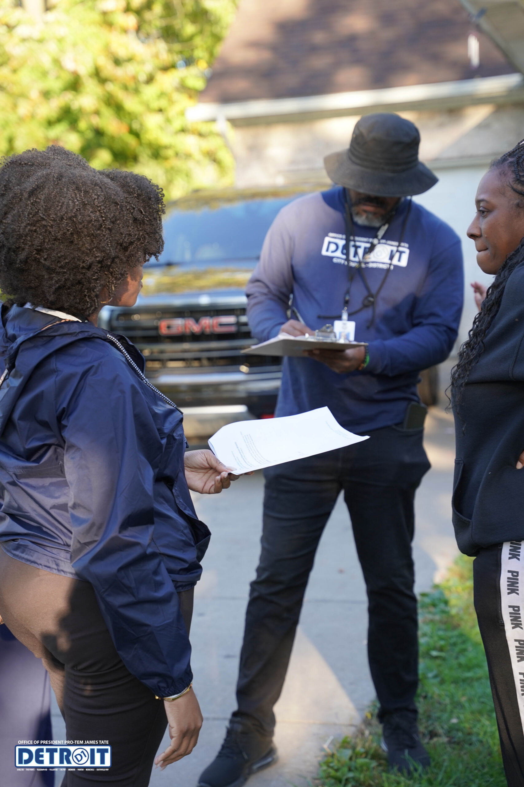 Two people speak while a man in a DETROIT shirt writes on a clipboard outdoors. A GMC vehicle is parked behind them, and all appear to be engaged in conversation or a survey on a residential street.