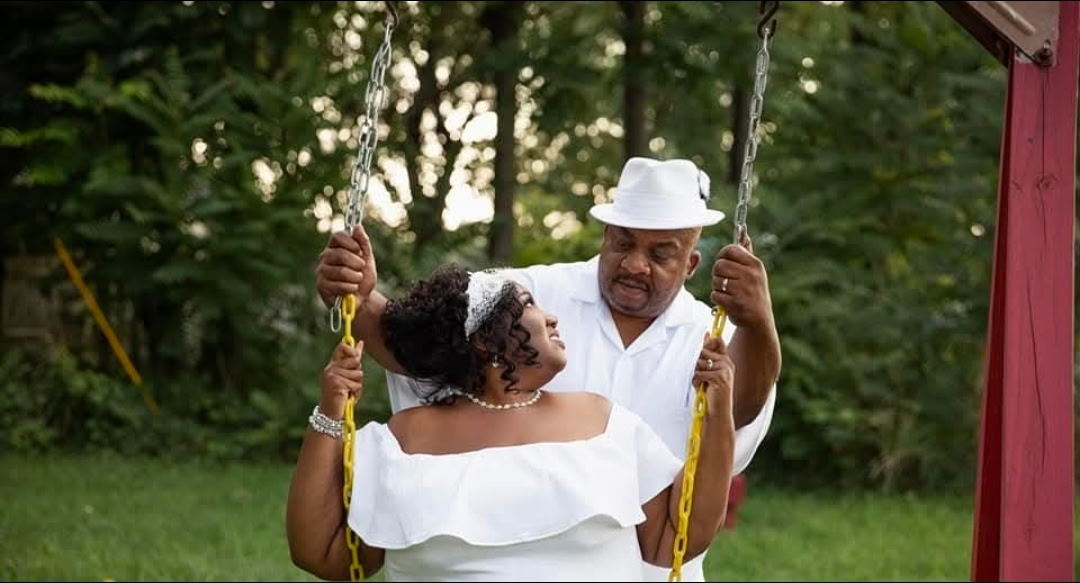 A couple dressed in white, with the woman sitting on a swing and the man standing behind her holding the chains, smiles at each other in a grassy outdoor setting surrounded by trees.