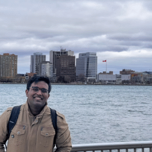A man in a tan jacket and glasses stands smiling by a riverside railing, with city buildings and a Canadian flag visible across the water under a cloudy sky.
