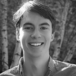 A young person with short dark hair smiles at the camera, wearing a checkered shirt. The background shows blurred tree trunks and foliage. The image is in black and white.