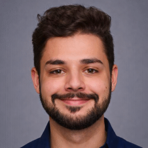 A young man with short, wavy dark hair, a beard, and mustache smiles at the camera. He is wearing a dark blue collared shirt and is posed against a plain gray background.