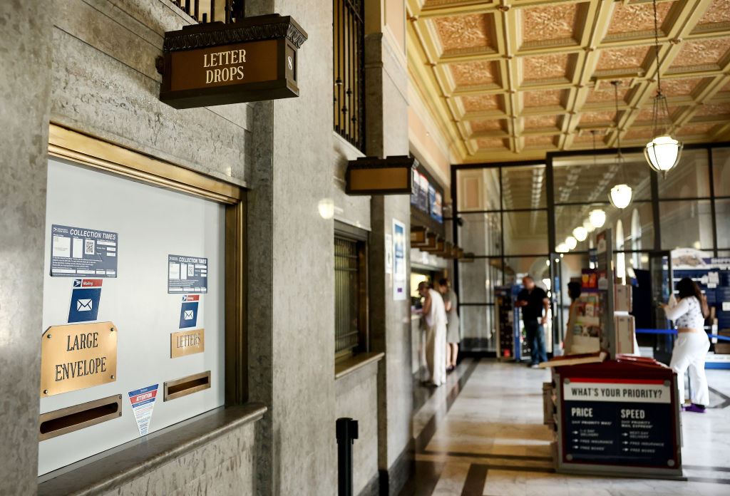 Interior of a post office with marble walls and a decorative ceiling, showing letter drop slots and a “WHAT’S YOUR PRIORITY?” postal information stand; people are waiting in line in the background.