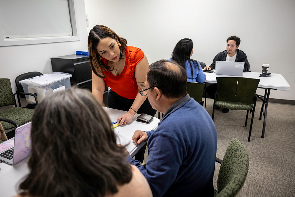 A woman in an orange top assists two seated people at a table while another pair works at a separate table in the background; all are in an office or classroom setting.