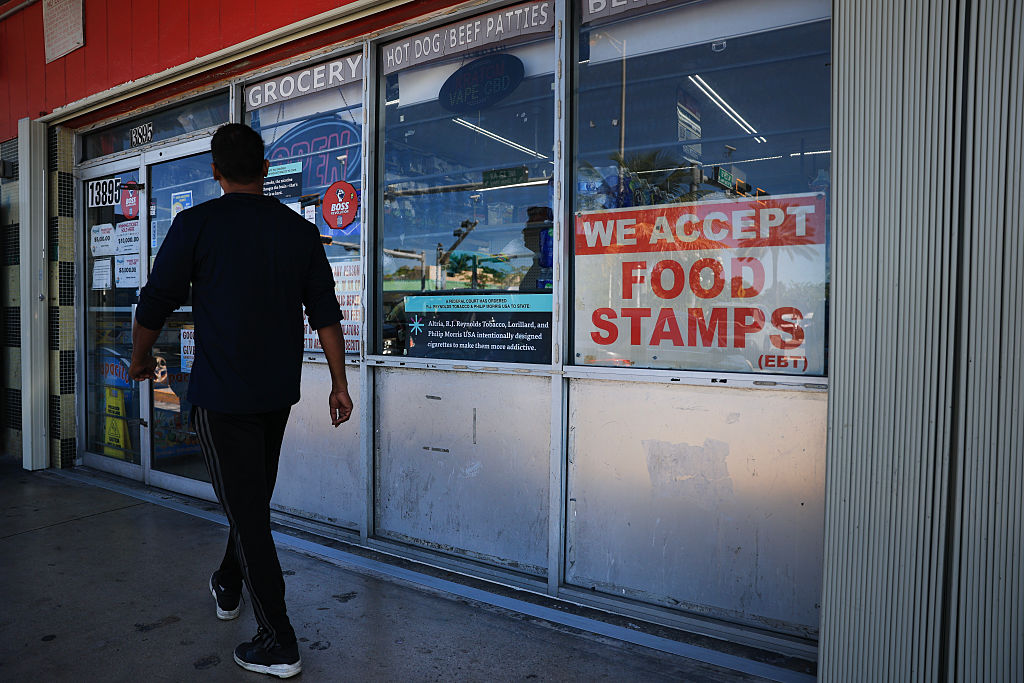 A person walks toward a store entrance with various signs on the glass, including a large one that reads WE ACCEPT FOOD STAMPS (EBT).