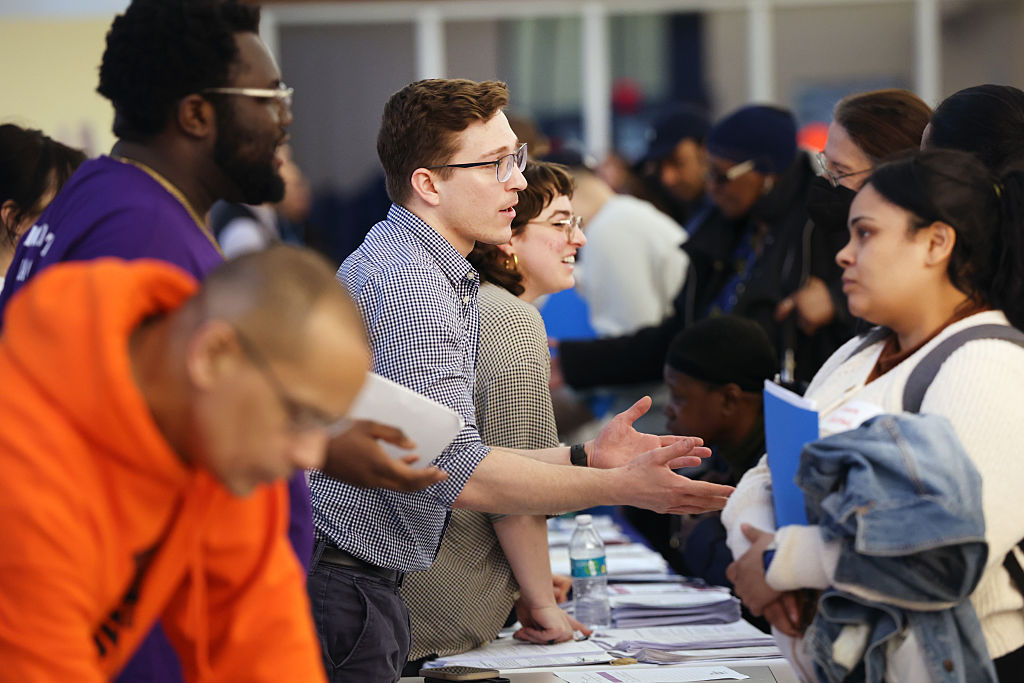 People interact across tables at a busy event, with one man in glasses and a checkered shirt talking to a woman holding documents. Other attendees and staff are visible in the background.