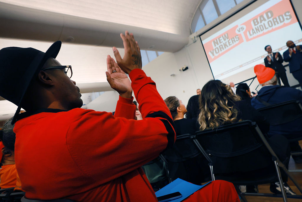 A person in a red outfit claps while seated among a crowd at an indoor event. A large screen in the background reads NEW YORKERS vs BAD LANDLORDS as speakers address the audience from a stage.
