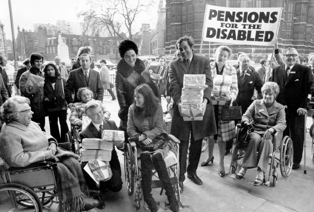 A group of people, some in wheelchairs, gather outdoors holding packages. Behind them, others stand and smile. A large banner in the background reads, PENSIONS FOR THE DISABLED.