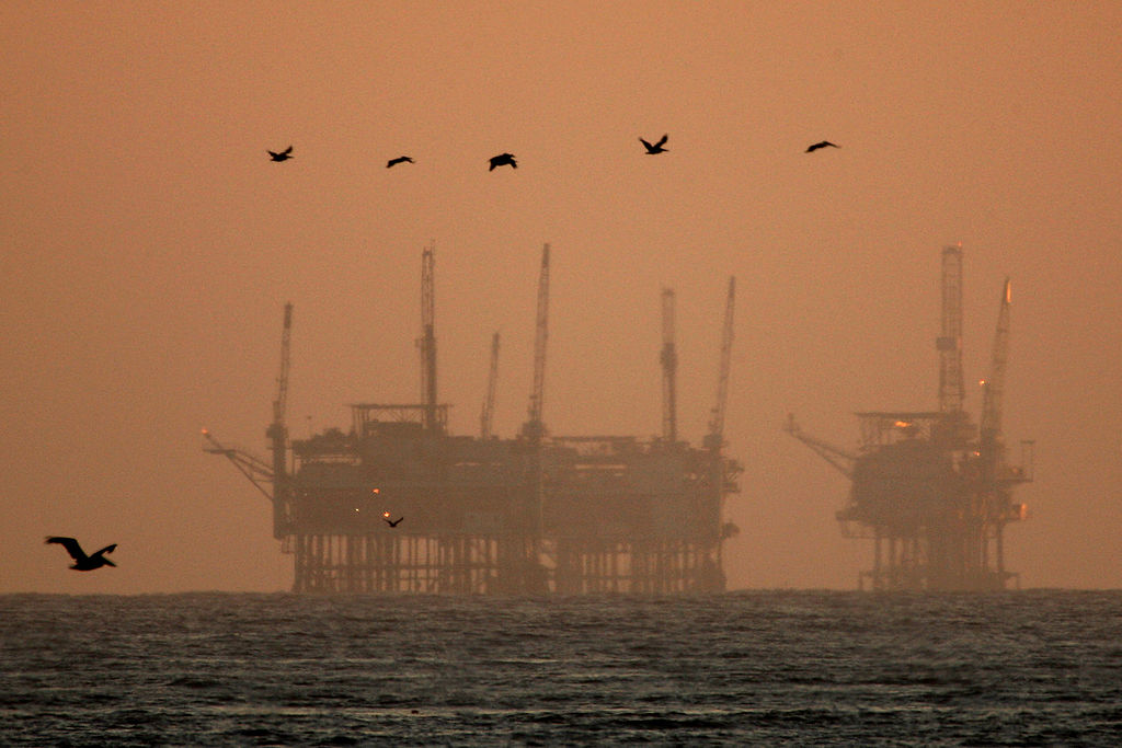 Offshore oil platforms stand in the ocean at sunset, silhouetted against an orange sky. Several birds are flying across the scene above the water.