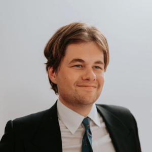 A young man with short brown hair, wearing a dark suit, white shirt, and patterned tie, smiles while looking slightly to the side against a plain light background.