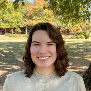 A woman with shoulder-length curly brown hair, wearing a light beige sweater and a gold necklace, smiles while standing outdoors in a park with trees and grass under daylight.