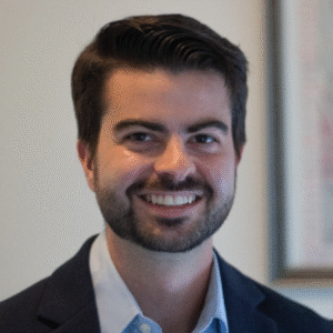 A man with dark hair and a beard, wearing a navy blazer and light blue shirt, smiles at the camera in a softly lit room with a blurred artwork in the background.