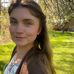 A young woman with long brown hair stands outdoors on a sunny day, smiling slightly. She is in a grassy area with trees and a wooden building in the background. Sunlight filters through the branches.