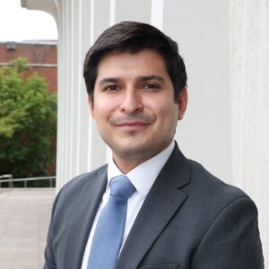 A man in a dark suit, white shirt, and light blue tie stands outdoors in front of a modern building with white columns. He has short dark hair and is smiling at the camera. Green trees and a brick building are in the background.