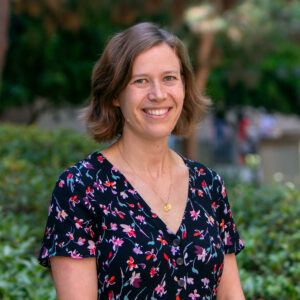 A woman with short brown hair wearing a floral dress and gold necklace stands outdoors, smiling at the camera. Green bushes and trees blur in the background.