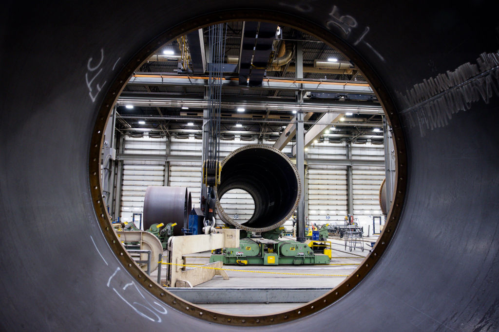 View through a large metal pipe inside a factory, with workers and machinery visible in the background and another large pipe aligned in the center, creating a tunnel effect.