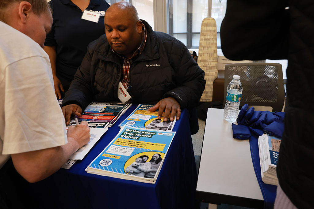 A man sits at a table handing out informational brochures. Another person is filling out a form. A water bottle, pens, and more pamphlets are on the table. The setting appears to be an indoor event or resource fair.