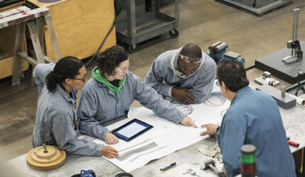 Four people in work uniforms gather around a table with blueprints and technical tools, discussing a project; one person uses a tablet, others look at documents, in an industrial workshop setting.