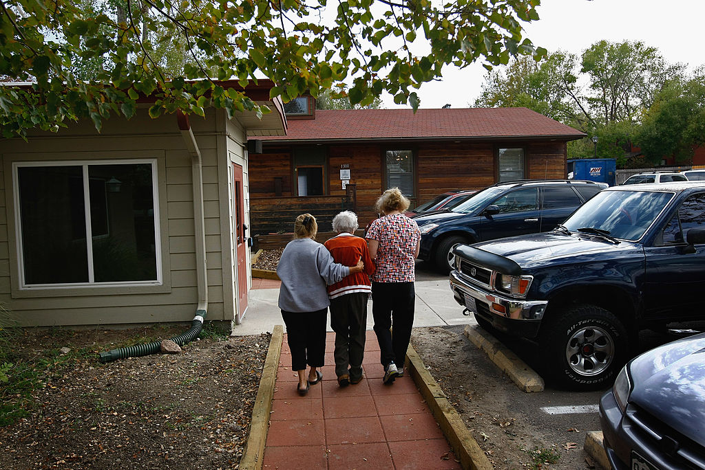 Three women walk arm in arm along a sidewalk toward a building, surrounded by parked cars and trees. The two women on either side support the woman in the middle.