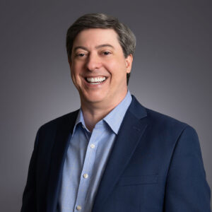 A man with short dark hair, wearing a blue suit jacket and light blue dress shirt, is smiling broadly in front of a plain dark gray background.