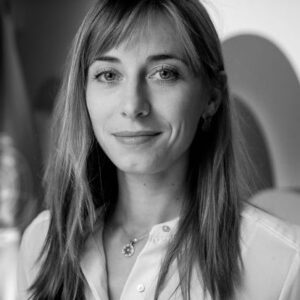 Black and white portrait of a young woman with straight, long hair, wearing a light-colored blouse and a necklace, looking at the camera with a slight smile. The background is softly blurred.
