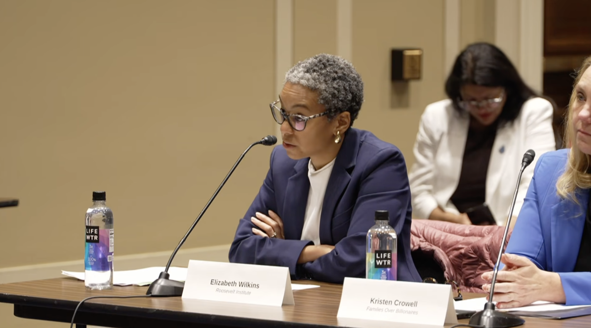 A woman in a blue blazer and glasses sits at a table with a microphone, speaking at a panel discussion. Two water bottles and nameplates labeled Elizabeth Wilkins and Kristen Crowell are visible. Others sit in the background.