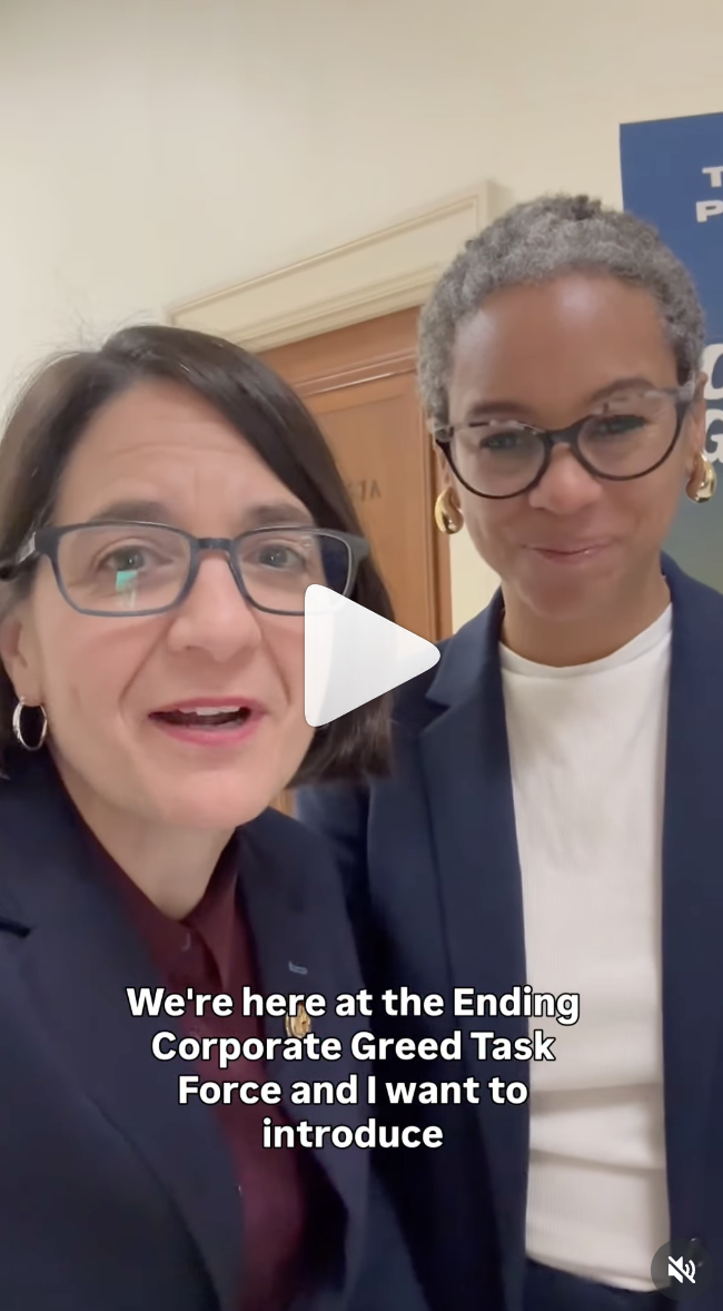 Two women in business attire stand together smiling indoors. The woman on the left is speaking, and white text at the bottom reads, Were here at the Ending Corporate Greed Task Force and I want to introduce.