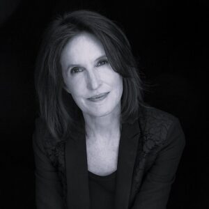 Katrina vanden Heuvel with shoulder-length dark hair and a slight smile looks at the camera. She wears a dark blazer with lace details on the shoulders, against a plain black background. The image is in black and white.