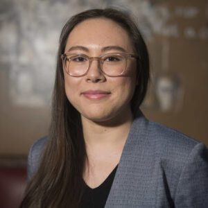 A person with long brown hair, wearing glasses, a light blue blazer, and a black top, stands in front of a blurred brown and gray background, looking at the camera and smiling slightly.