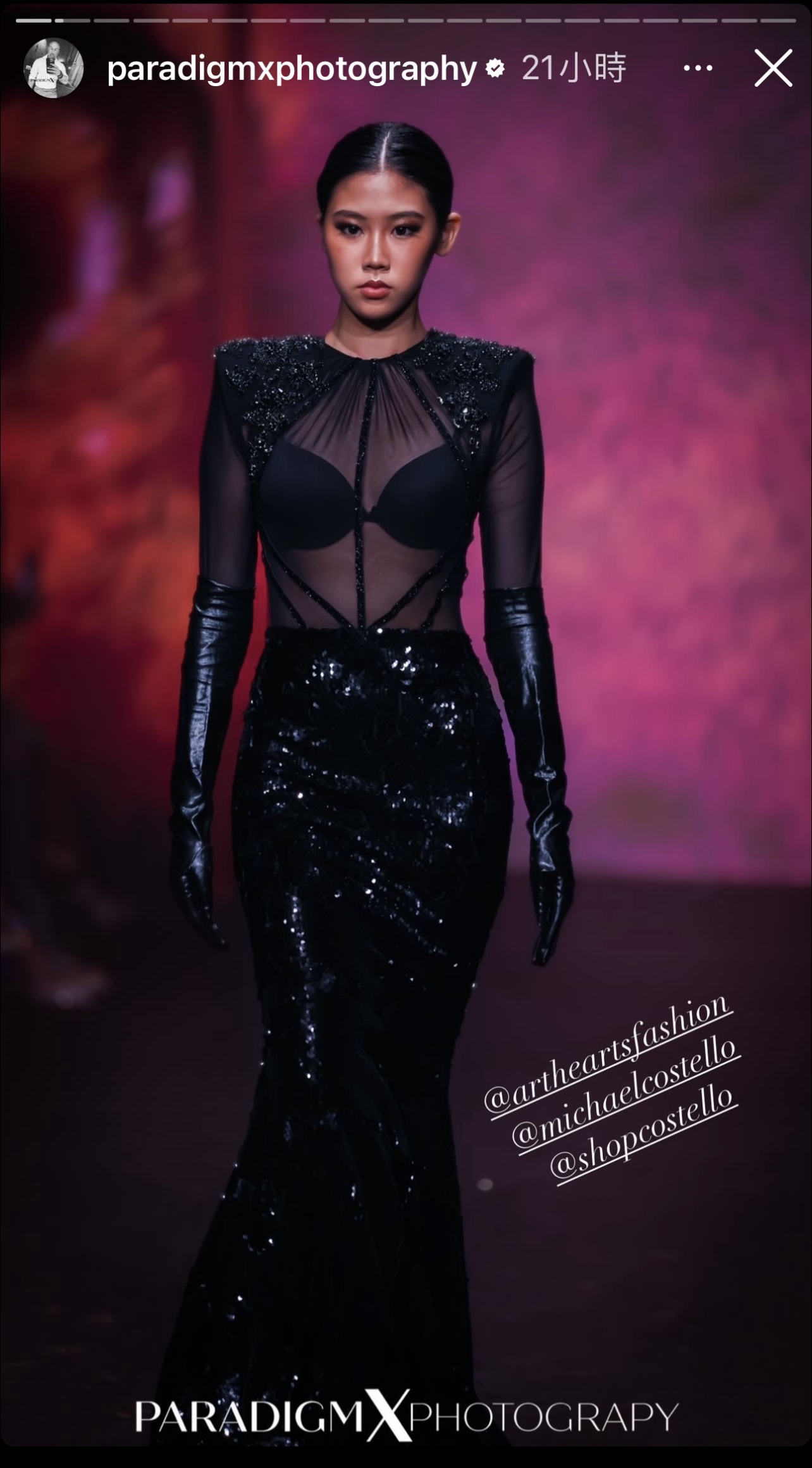 A model walks the runway in a black sequin gown with sheer panels, structured shoulders, and long black gloves. The background is dark with reddish lighting, giving a dramatic effect.