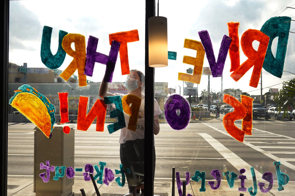 A person paints a colorful sign reading DRIVE-THRU $0 FEE! with a taco illustration on a restaurant window facing a busy street. Sunlight and cars are visible outside, and the painter wears a mask.