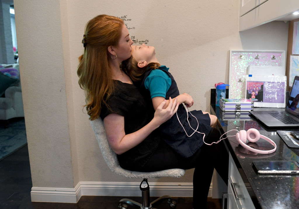 A woman sits at a desk holding a young girl leaning back in her lap. The woman looks lovingly at the child. On the desk are a laptop, a notebook, and pink headphones. A sofa is visible in the background.