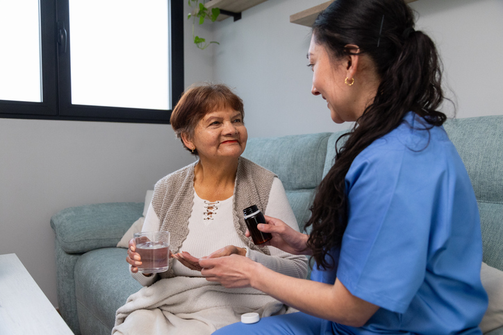A nurse in blue scrubs hands a bottle of medication to an elderly woman sitting on a couch, who is holding a glass of water and smiling warmly at the nurse.