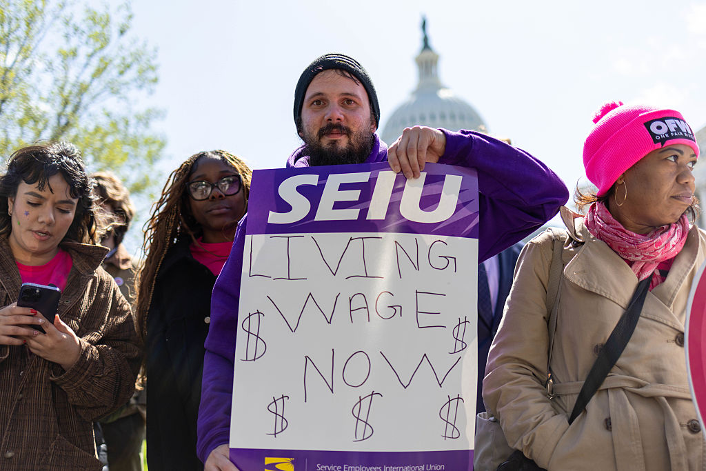 A man holds a sign reading SEIU LIVING WAGE NOW with dollar signs, standing among a group of people at a protest. The U.S. Capitol building is visible in the background.