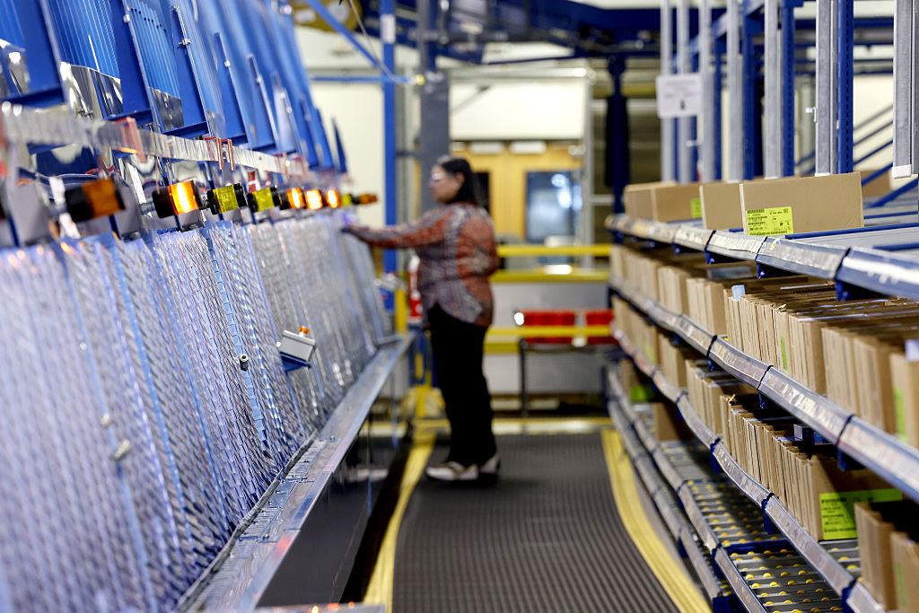 A person stands in a warehouse aisle, operating sorting machinery with shelves of packages on both sides, indicating a parcel sorting or distribution center environment.