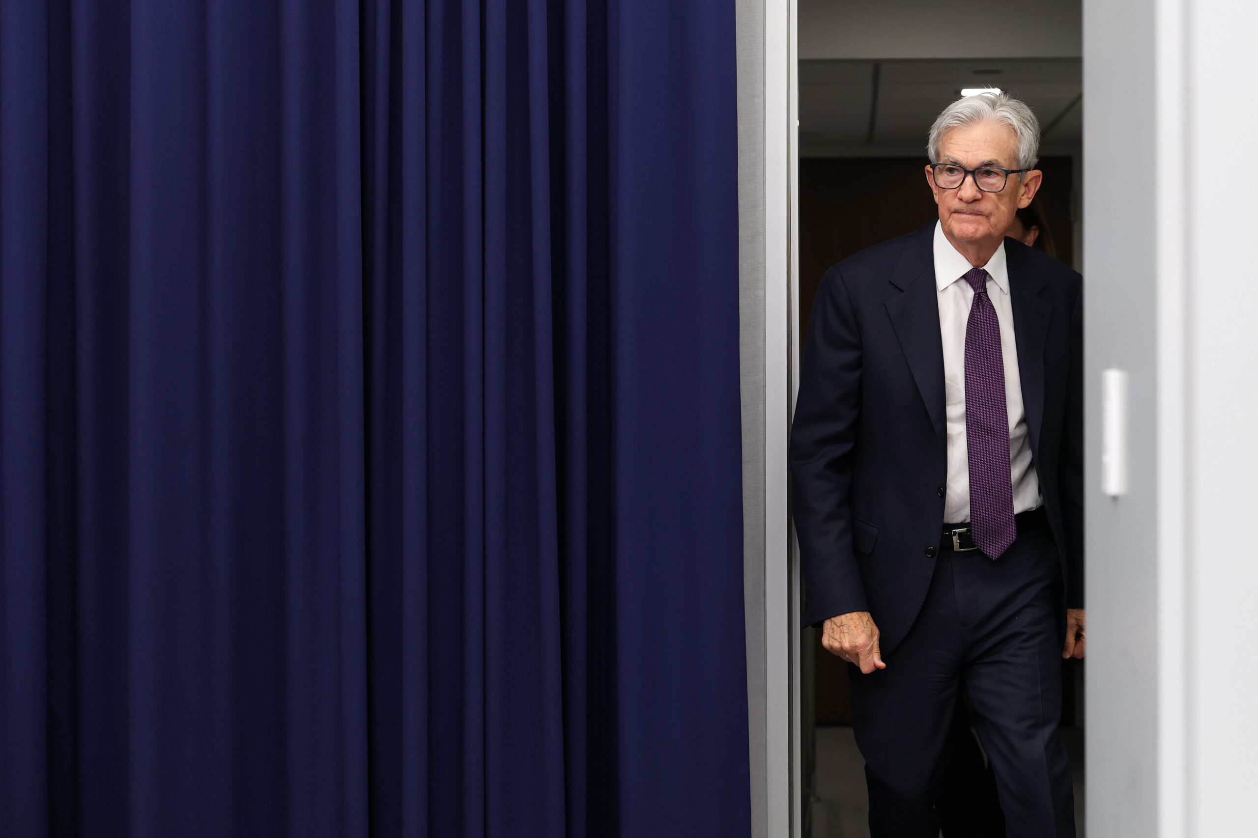A man in a dark suit, white shirt, and purple tie walks through a doorway with blue curtains on the left side, entering a room with gray walls.