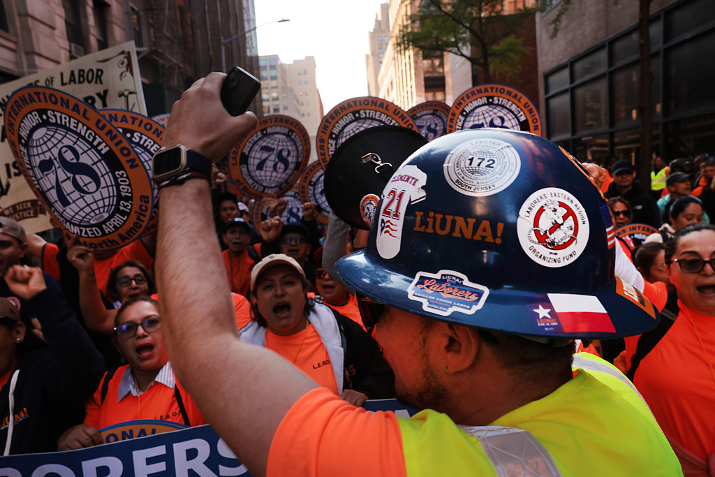 A construction worker wearing a sticker-covered hard hat raises his fist while leading a group of people in bright orange shirts at a city protest or rally. Signs and banners are visible in the background.