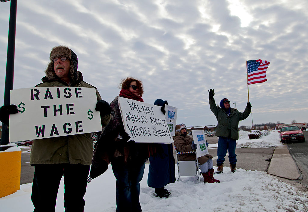Four people stand and sit in the snow during a protest. Two hold signs reading RAISE THE WAGE and Walmart Americas Biggest Welfare Queen! Another holds an American flag, and cloudy sky is visible above.