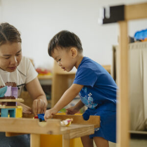 An adult and a young child play with building blocks at a wooden table in a brightly lit room, focusing on assembling pieces together. Toys and storage bins are visible in the background.